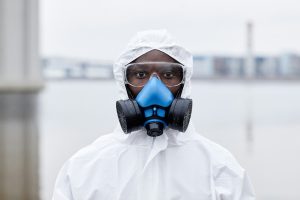 A person wearing a white protective suit, blue respiratory mask, and clear goggles stands outdoors. The background is blurred but shows an industrial or urban setting near water, indicating the effectiveness of interventions by Toxic Teams in hazardous environments.