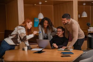 A group of four people gather around a laptop on a wooden table in a modern workspace. They are smiling and engaged, with one person pointing at the laptop screen. Books, a cup, and a notebook are also on the table—hallmarks of effective interventions in team management. No toxic teams here!